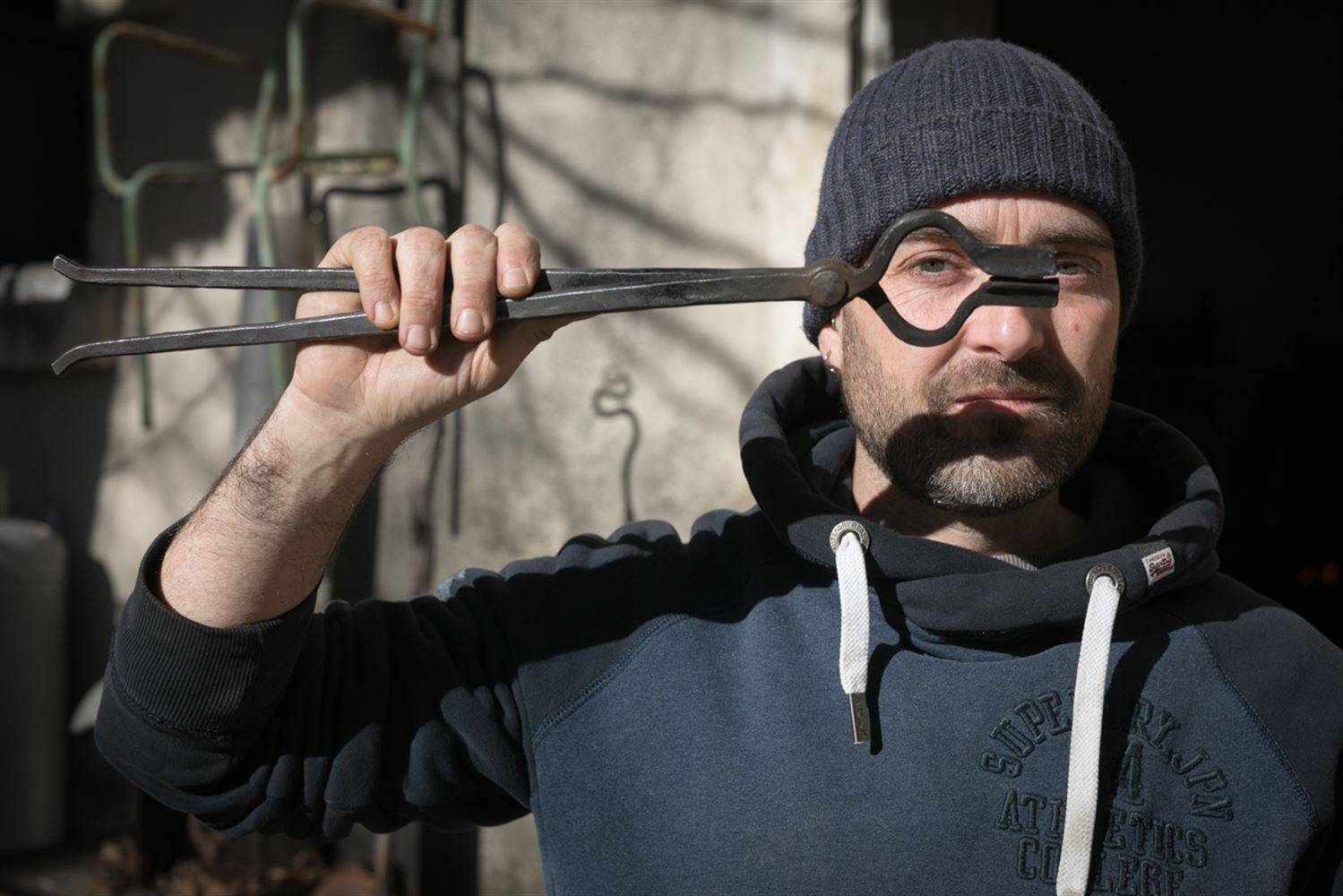 Dans la cour devant son atelier-forge, Julien tient une tenaille de forgeron devant son oeil droit que l'on voit à travers le mors à demi-ronds.